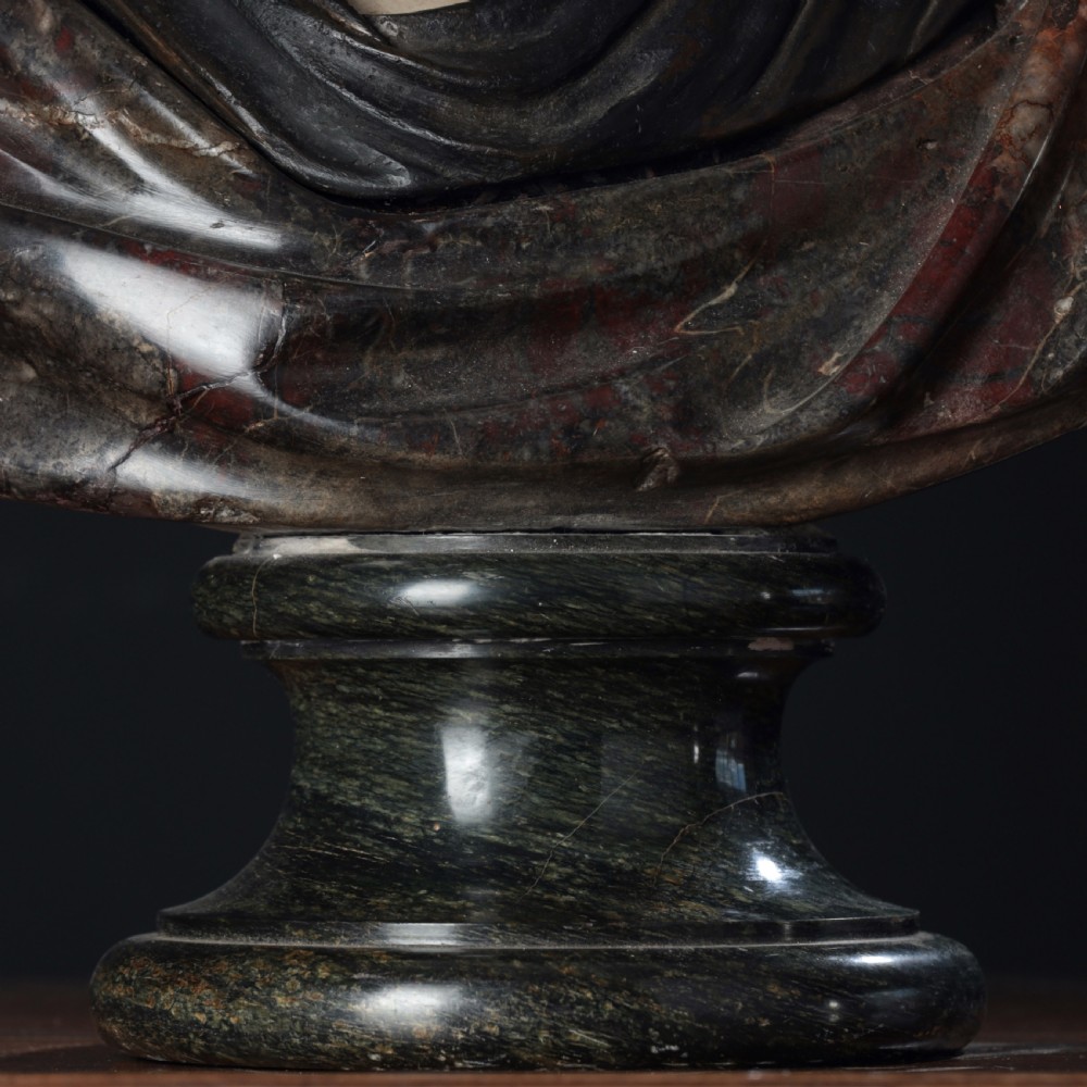 A carved marble profile bust depicting a woman with wavy hair gathered in a bun, wearing dark draped robes, set upon a turned marble socle. View IX