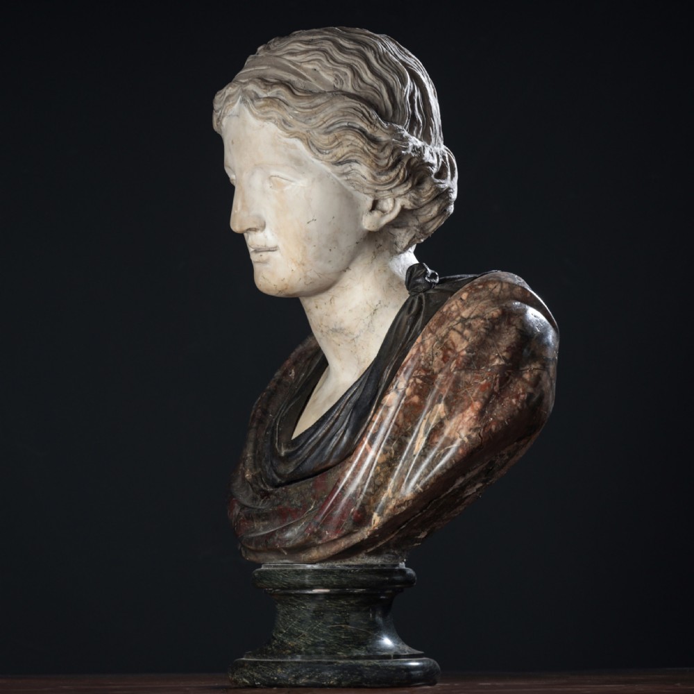 A carved marble profile bust depicting a woman with wavy hair gathered in a bun, wearing dark draped robes, set upon a turned marble socle. View X