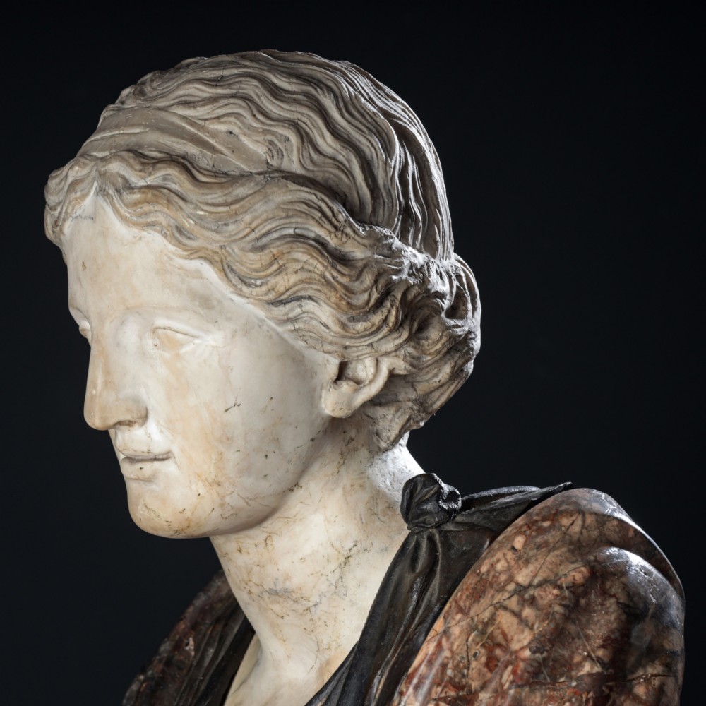 A carved marble profile bust depicting a woman with wavy hair gathered in a bun, wearing dark draped robes, set upon a turned marble socle. View XI