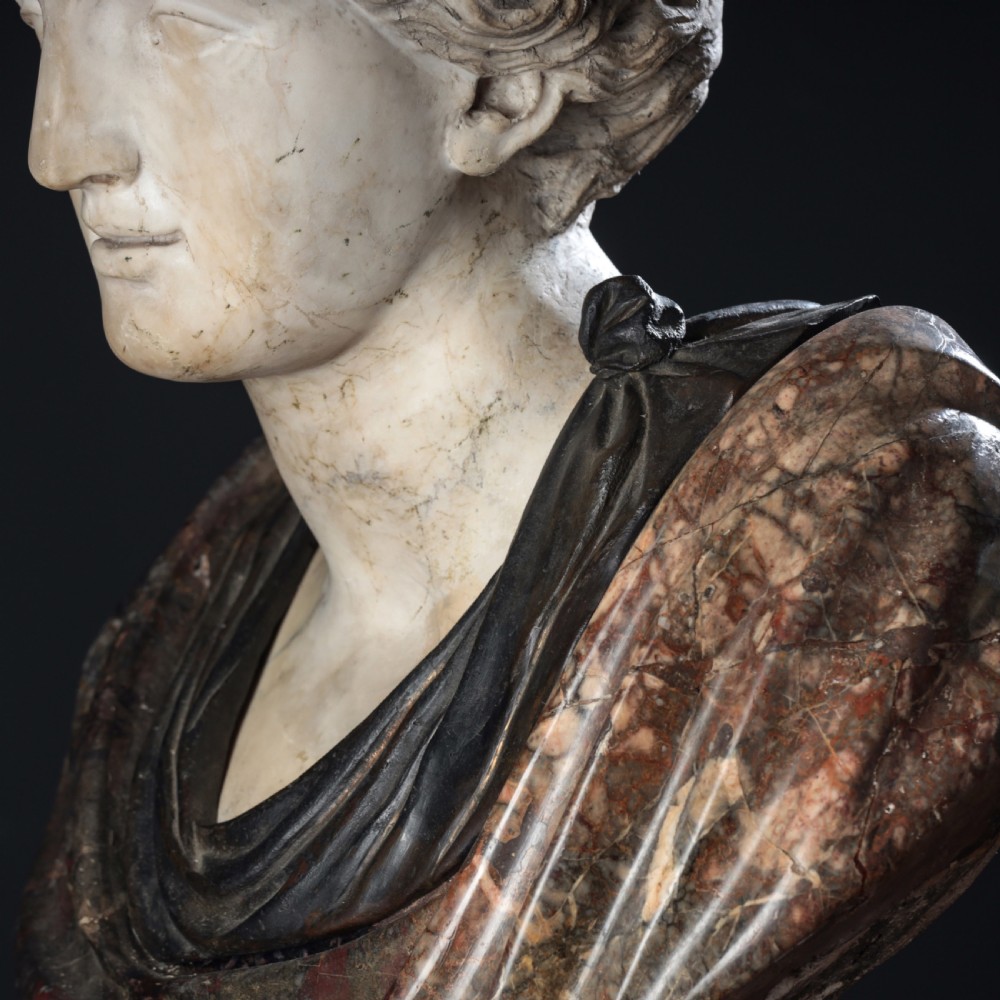 A carved marble profile bust depicting a woman with wavy hair gathered in a bun, wearing dark draped robes, set upon a turned marble socle. View XII