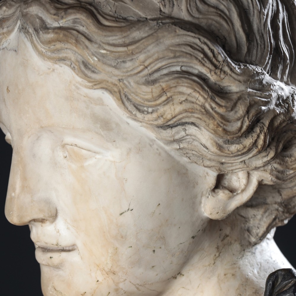 A carved marble profile bust depicting a woman with wavy hair gathered in a bun, wearing dark draped robes, set upon a turned marble socle. View XIII