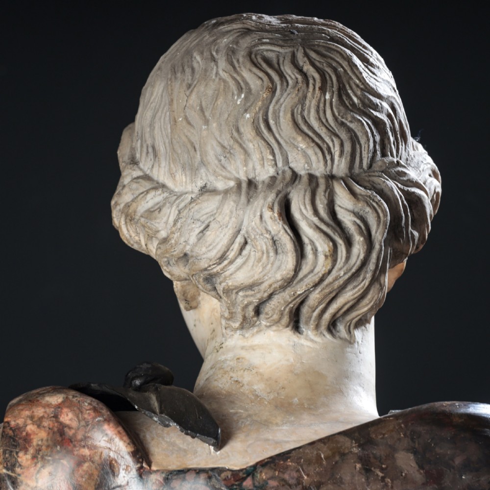 A carved marble profile bust depicting a woman with wavy hair gathered in a bun, wearing dark draped robes, set upon a turned marble socle. View XV
