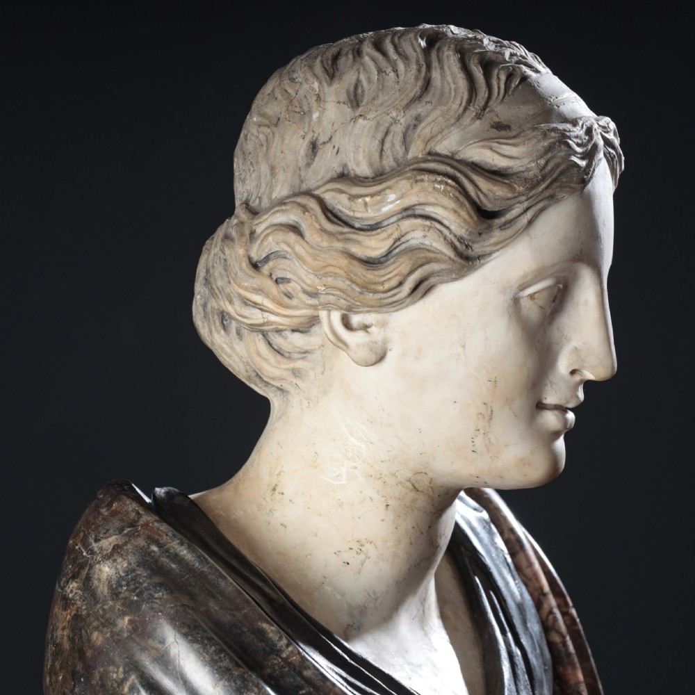 A carved marble profile bust depicting a woman with wavy hair gathered in a bun, wearing dark draped robes, set upon a turned marble socle. View XVIII