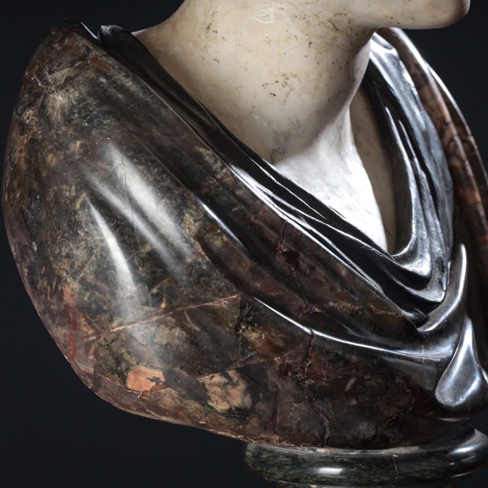 A carved marble profile bust depicting a woman with wavy hair gathered in a bun, wearing dark draped robes, set upon a turned marble socle. View XIX