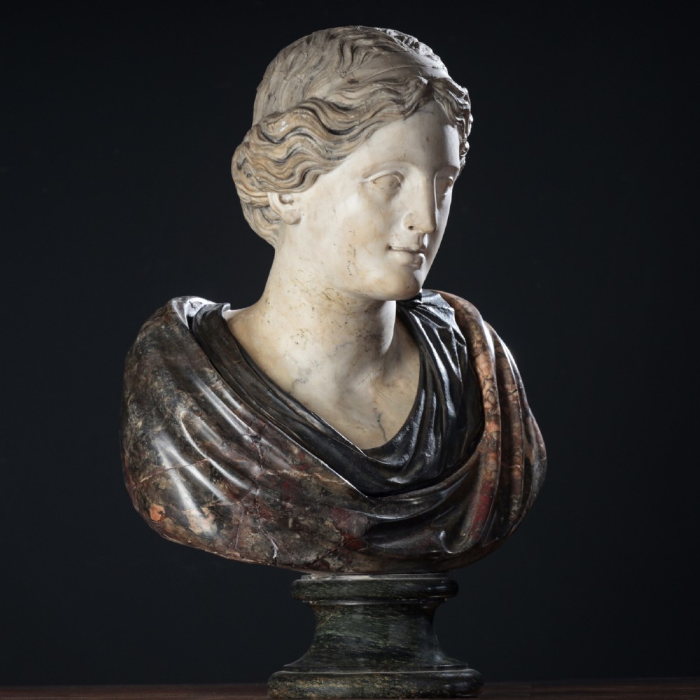 A carved marble profile bust depicting a woman with wavy hair gathered in a bun, wearing dark draped robes, set upon a turned marble socle. View XX