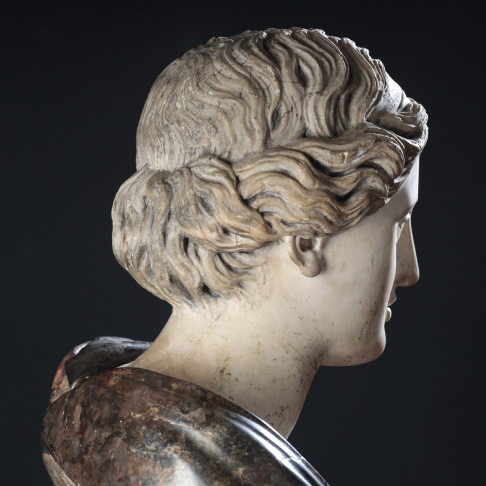 A carved marble profile bust depicting a woman with wavy hair gathered in a bun, wearing dark draped robes, set upon a turned marble socle. View XXI
