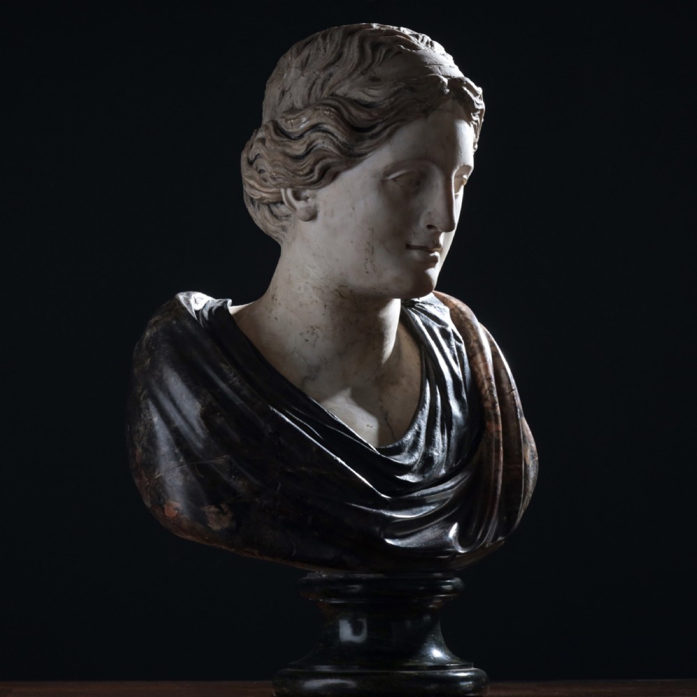A carved marble profile bust depicting a woman with wavy hair gathered in a bun, wearing dark draped robes, set upon a turned marble socle. View XXIII