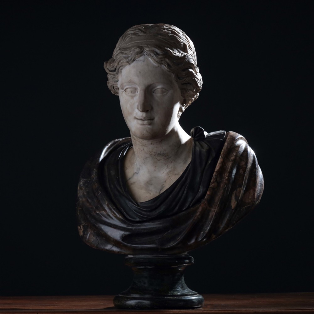 A carved marble profile bust depicting a woman with wavy hair gathered in a bun, wearing dark draped robes, set upon a turned marble socle. View XXIV