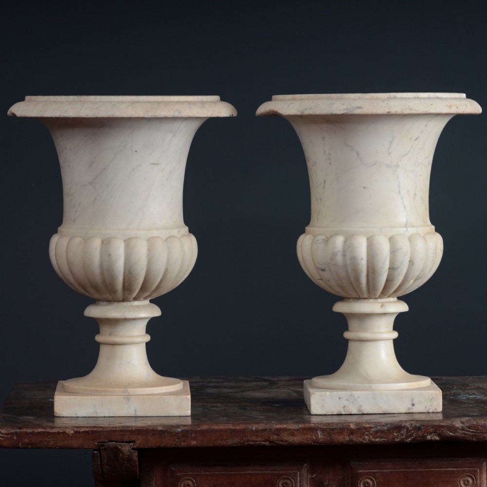 A pair of hand-carved white marble campana urns featuring gadrooned lower bodies and flared rims raised on square plinths.