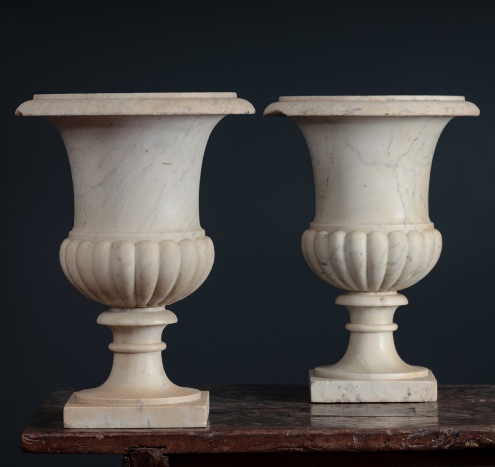 A pair of hand-carved white marble campana urns featuring gadrooned lower bodies and flared rims raised on square plinths. View V