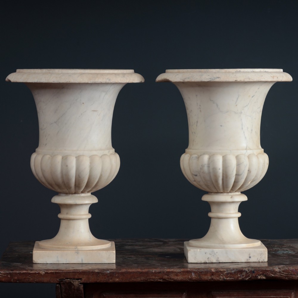 A pair of hand-carved white marble campana urns featuring gadrooned lower bodies and flared rims raised on square plinths. View VI