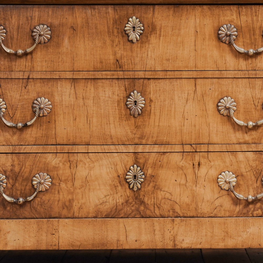An 18th-century walnut commode featuring three wide drawers decorated with brass rosette swing handles and keyhole escutcheons on tapered feet. View V