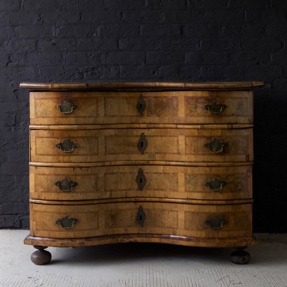 An 18th-century South German walnut chest of drawers featuring a serpentine front, four crossbanded drawers, brass drop handles, and turned bun feet.