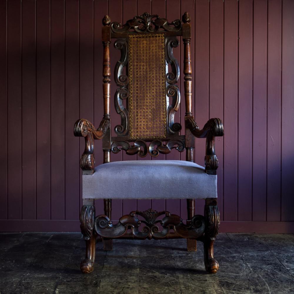 A 17th-century Carolean dark walnut armchair featuring a tall scroll-carved back with a central cane panel, scrolled armrests, and a grey upholstered seat.
