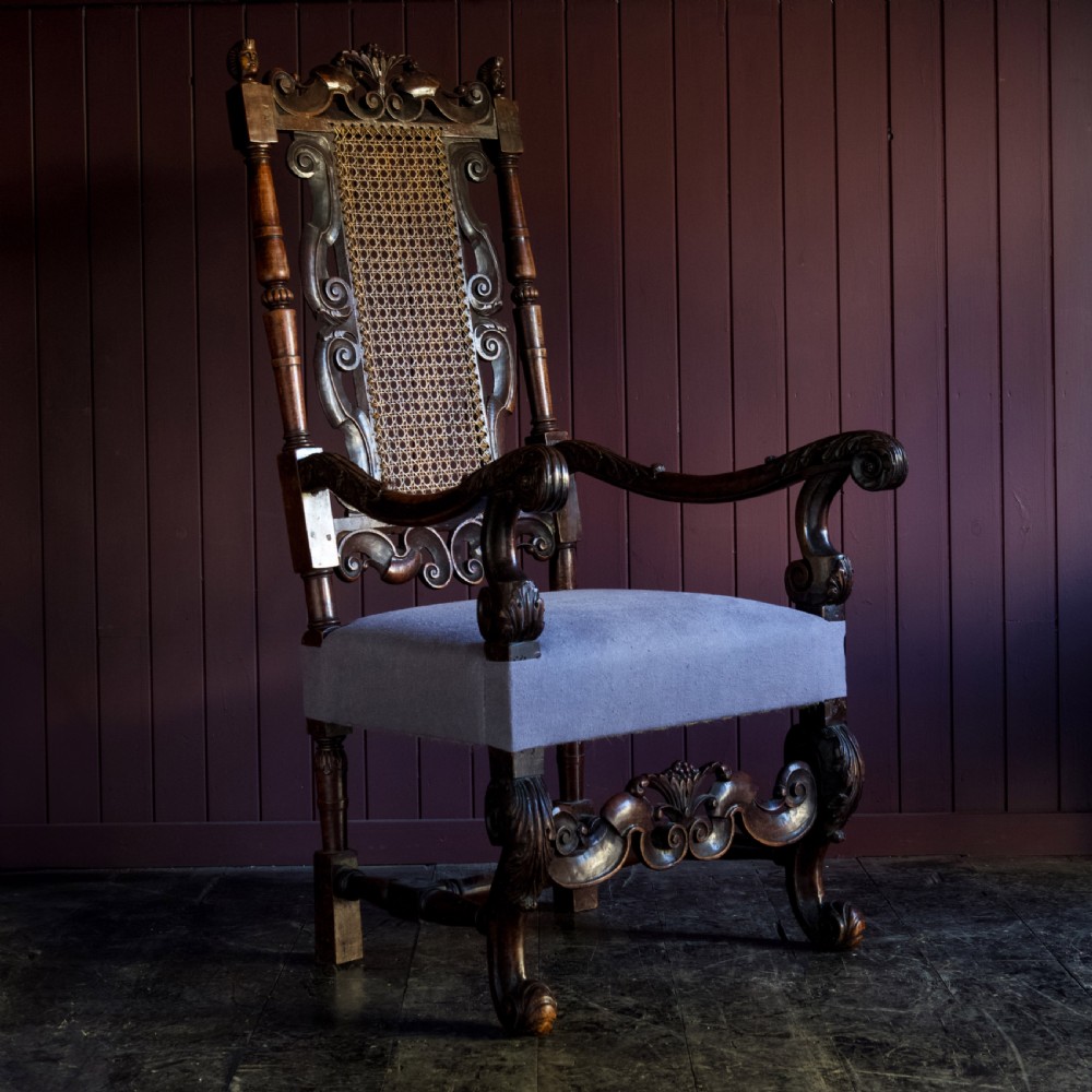 A 17th-century Carolean dark walnut armchair featuring a tall scroll-carved back with a central cane panel, scrolled armrests, and a grey upholstered seat. View II