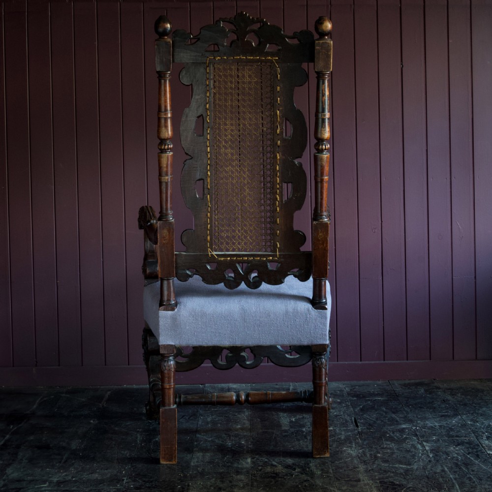 A 17th-century Carolean dark walnut armchair featuring a tall scroll-carved back with a central cane panel, scrolled armrests, and a grey upholstered seat. View III