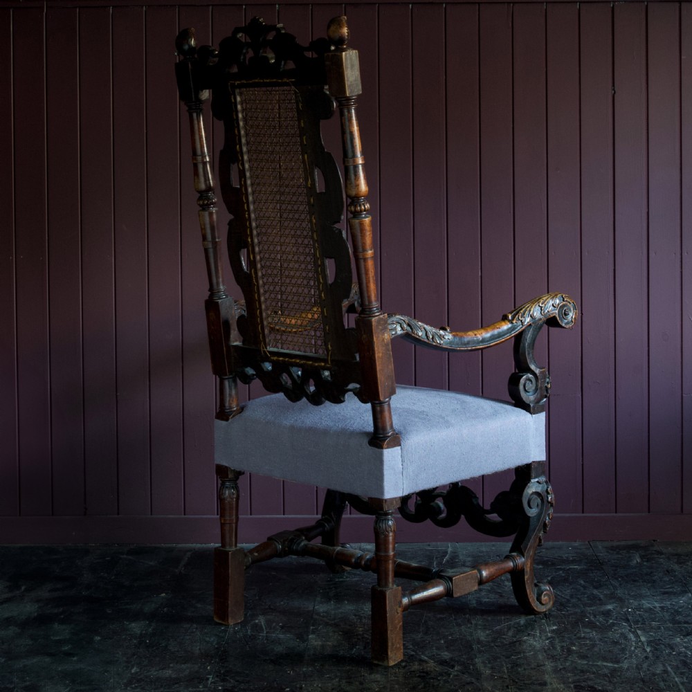 A 17th-century Carolean dark walnut armchair featuring a tall scroll-carved back with a central cane panel, scrolled armrests, and a grey upholstered seat. View IV