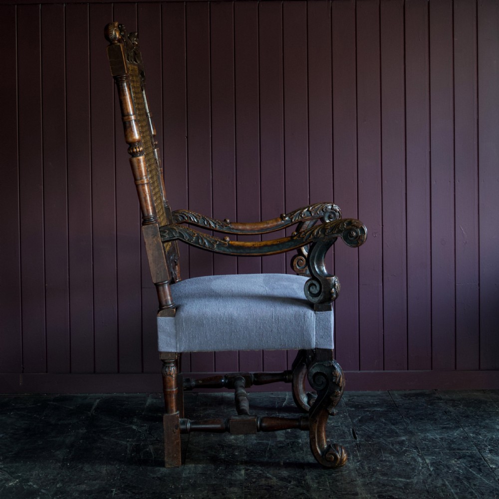 A 17th-century Carolean dark walnut armchair featuring a tall scroll-carved back with a central cane panel, scrolled armrests, and a grey upholstered seat. View V