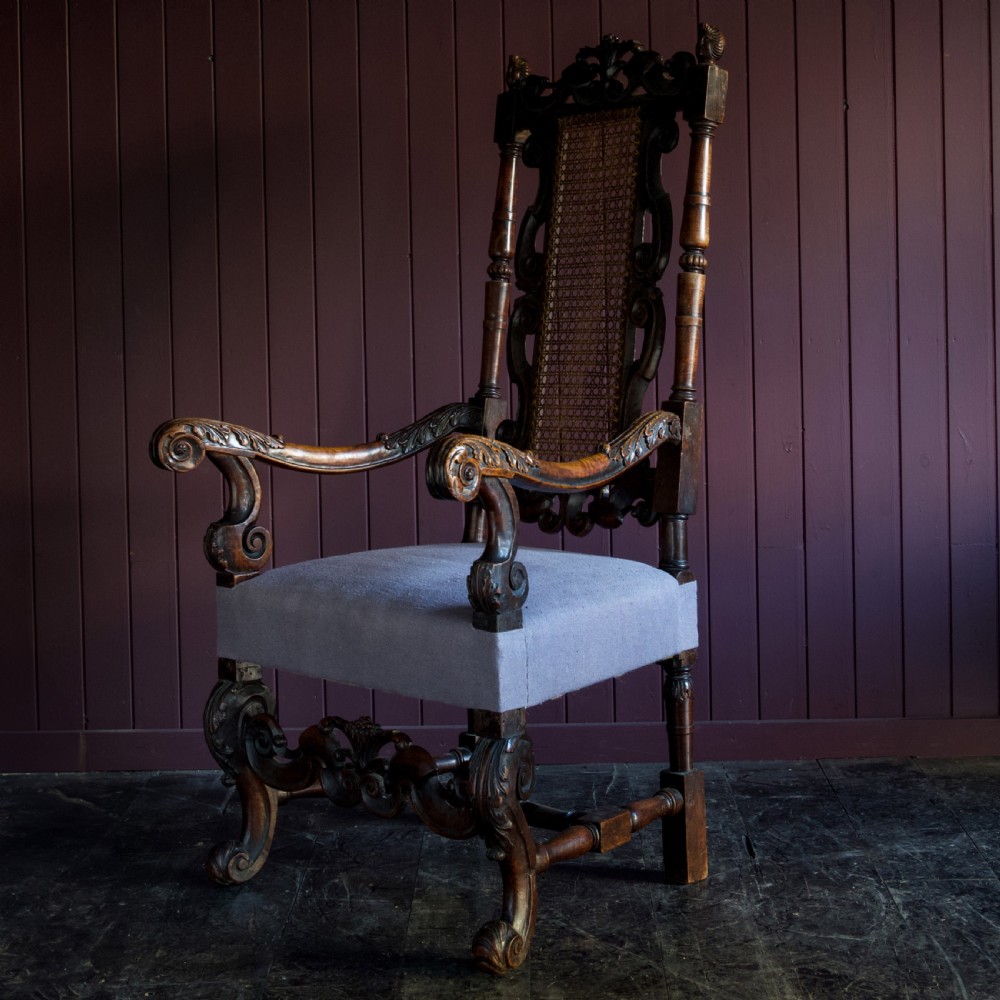 A 17th-century Carolean dark walnut armchair featuring a tall scroll-carved back with a central cane panel, scrolled armrests, and a grey upholstered seat. View VII