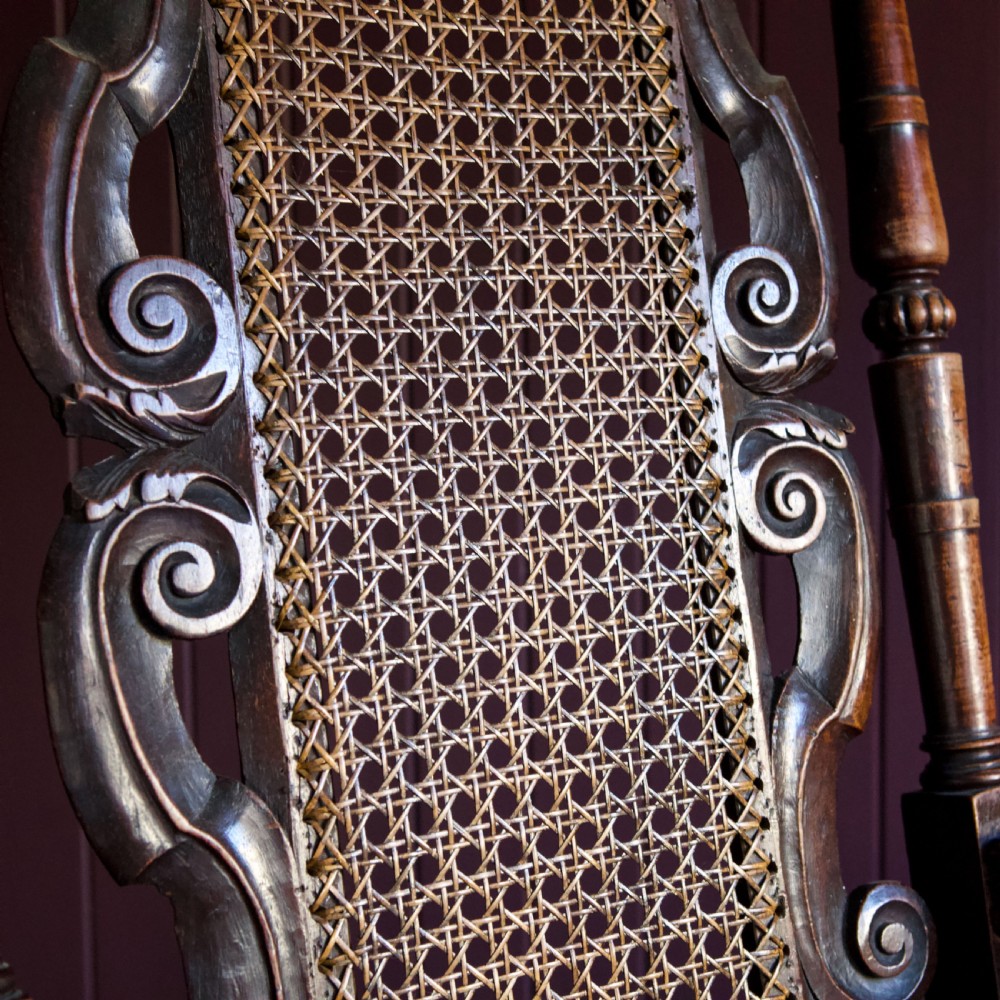A 17th-century Carolean dark walnut armchair featuring a tall scroll-carved back with a central cane panel, scrolled armrests, and a grey upholstered seat. View XI