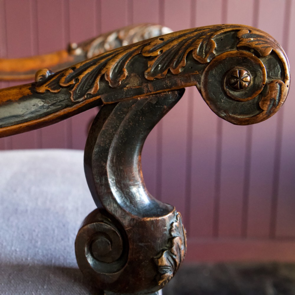 A 17th-century Carolean dark walnut armchair featuring a tall scroll-carved back with a central cane panel, scrolled armrests, and a grey upholstered seat. View XIII