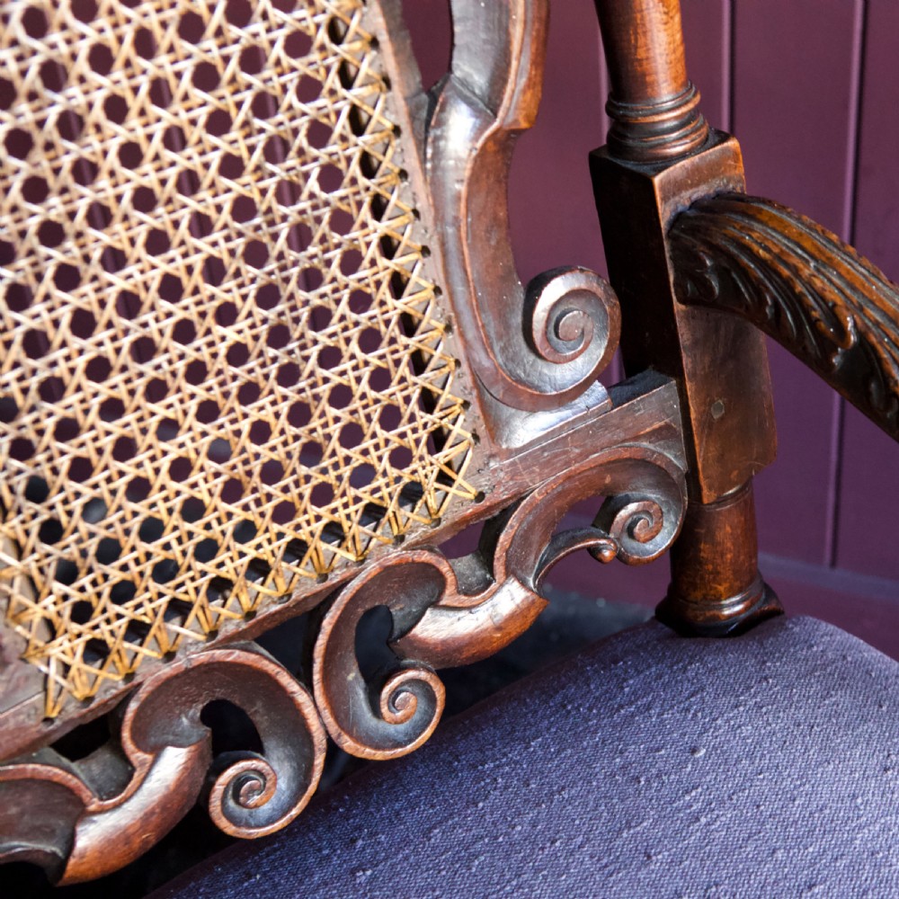 A 17th-century Carolean dark walnut armchair featuring a tall scroll-carved back with a central cane panel, scrolled armrests, and a grey upholstered seat. View XIV