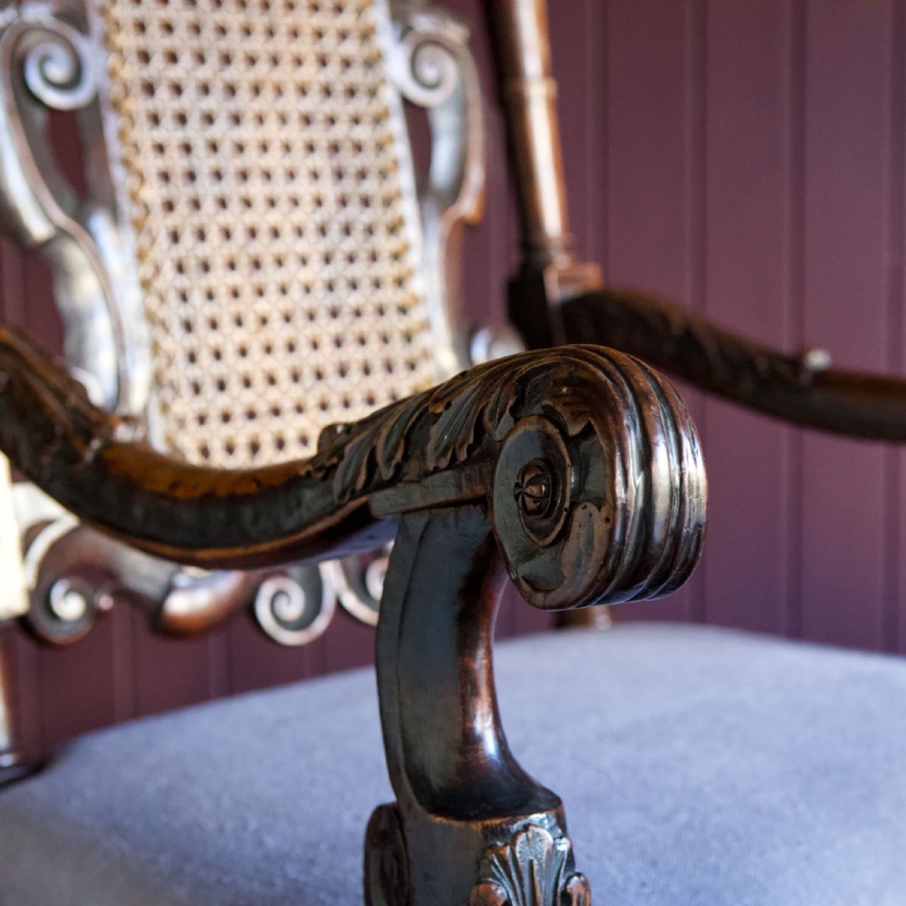A 17th-century Carolean dark walnut armchair featuring a tall scroll-carved back with a central cane panel, scrolled armrests, and a grey upholstered seat. View XV