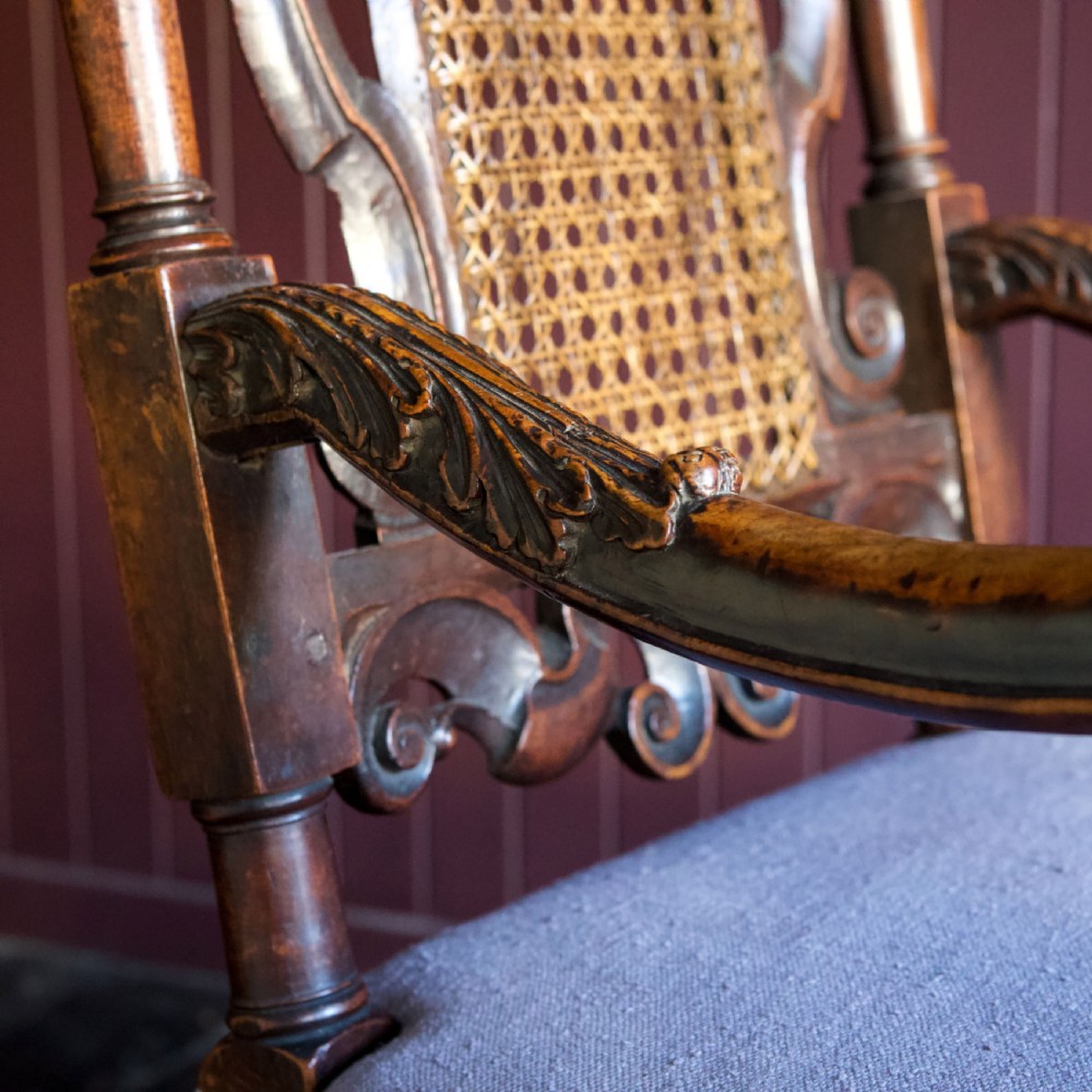 A 17th-century Carolean dark walnut armchair featuring a tall scroll-carved back with a central cane panel, scrolled armrests, and a grey upholstered seat. View XVI
