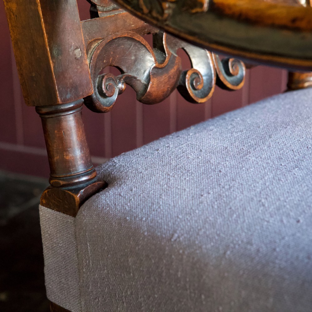 A 17th-century Carolean dark walnut armchair featuring a tall scroll-carved back with a central cane panel, scrolled armrests, and a grey upholstered seat. View XVII