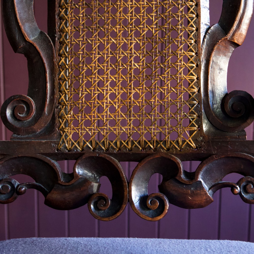 A 17th-century Carolean dark walnut armchair featuring a tall scroll-carved back with a central cane panel, scrolled armrests, and a grey upholstered seat. View XX