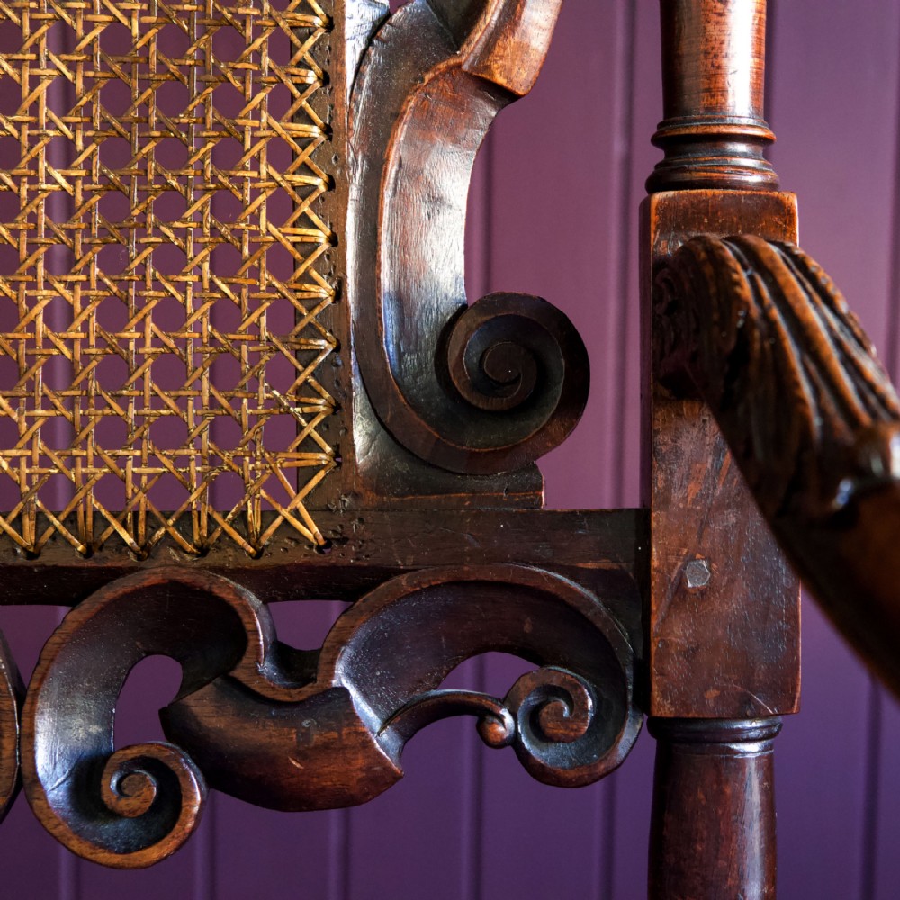 A 17th-century Carolean dark walnut armchair featuring a tall scroll-carved back with a central cane panel, scrolled armrests, and a grey upholstered seat. View XXI