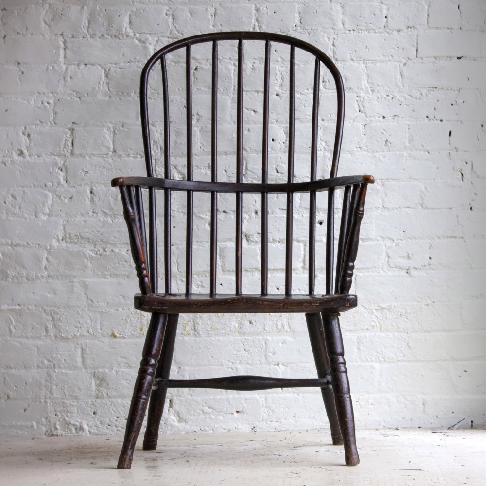 An 18th-century dark brown wooden stickback Windsor armchair featuring a tall curved hoop back, turned legs, and an H-stretcher.