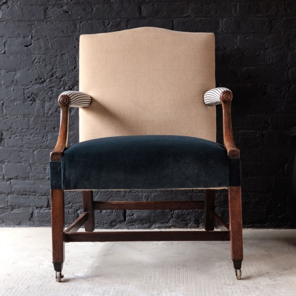 A George II Gainsborough armchair featuring an exposed timber frame, a high beige linen backrest, padded striped armrests, and a deep blue velvet upholstered seat on square legs with brass castors.