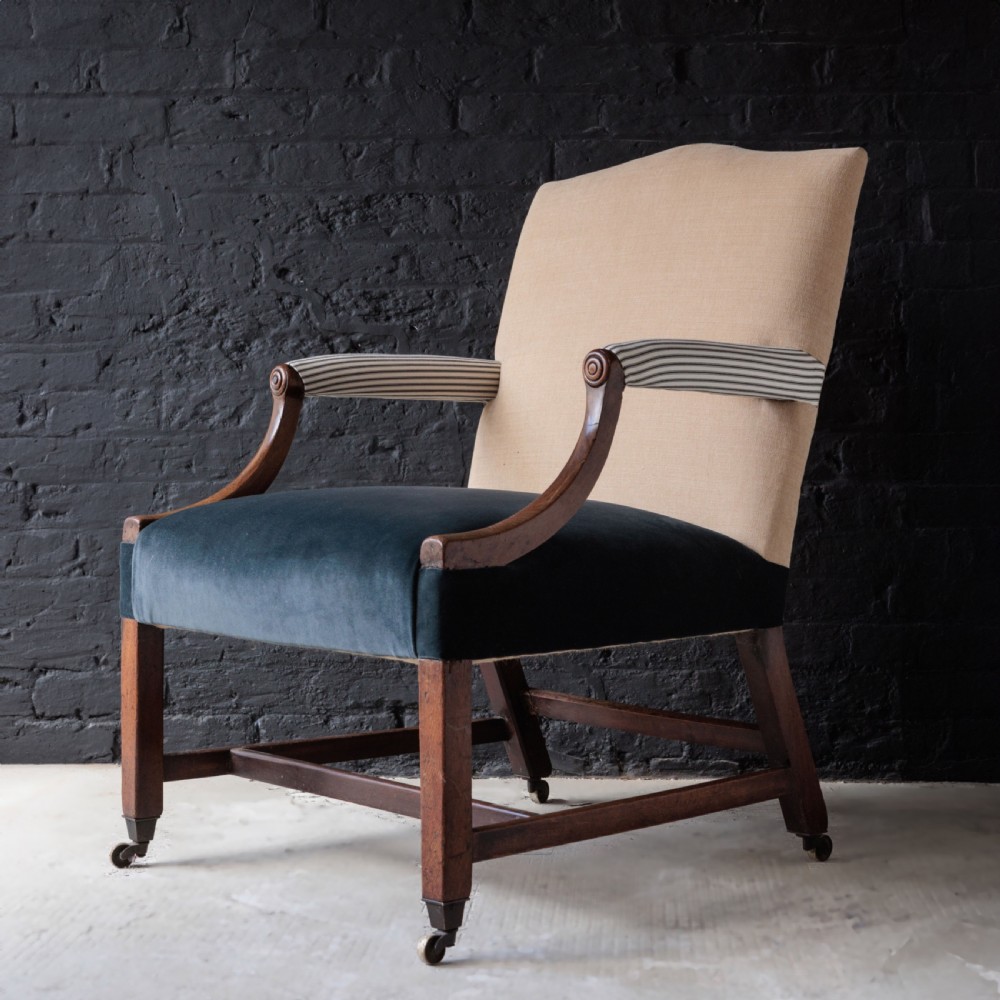 A George II Gainsborough armchair featuring an exposed timber frame, a high beige linen backrest, padded striped armrests, and a deep blue velvet upholstered seat on square legs with brass castors. View II