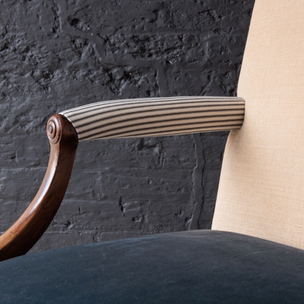 A George II Gainsborough armchair featuring an exposed timber frame, a high beige linen backrest, padded striped armrests, and a deep blue velvet upholstered seat on square legs with brass castors. View IV