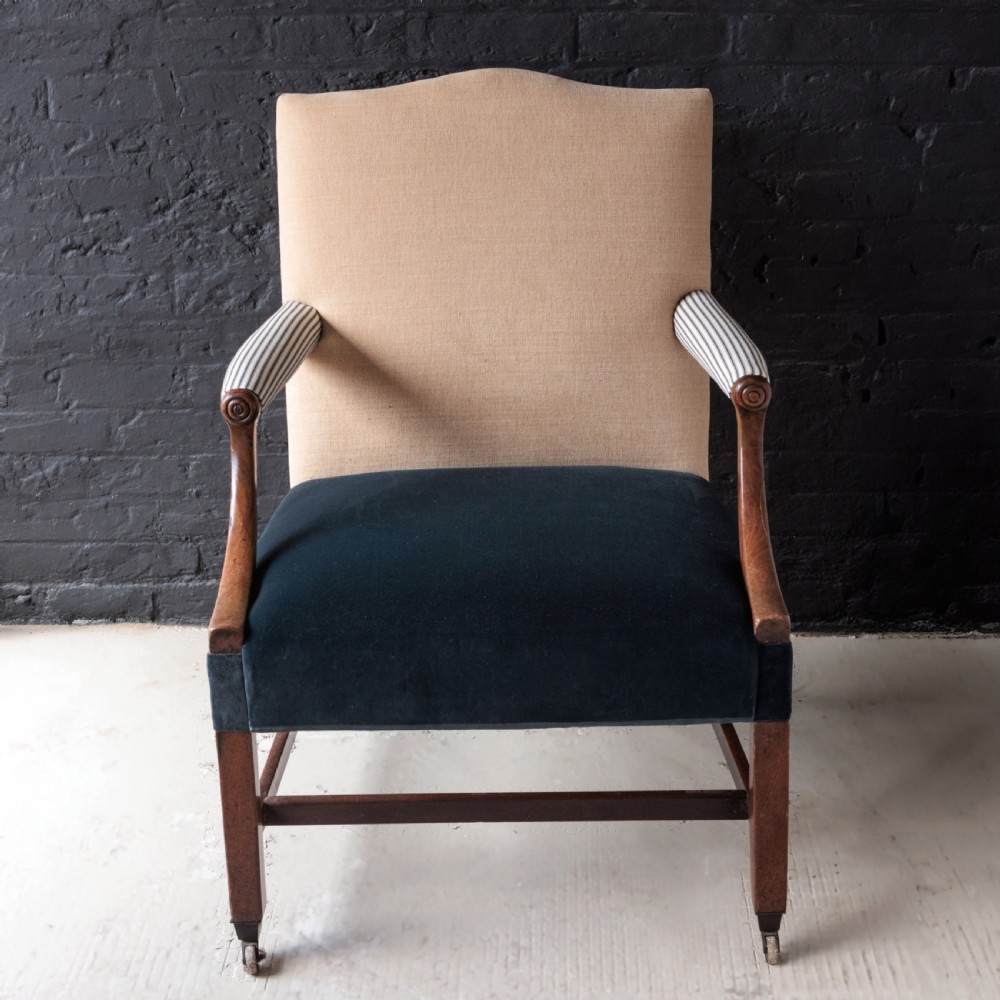 A George II Gainsborough armchair featuring an exposed timber frame, a high beige linen backrest, padded striped armrests, and a deep blue velvet upholstered seat on square legs with brass castors. View V