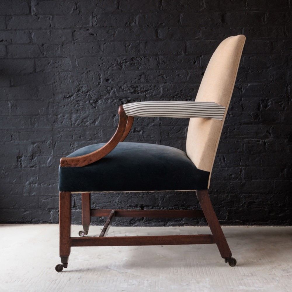 A George II Gainsborough armchair featuring an exposed timber frame, a high beige linen backrest, padded striped armrests, and a deep blue velvet upholstered seat on square legs with brass castors. View VII