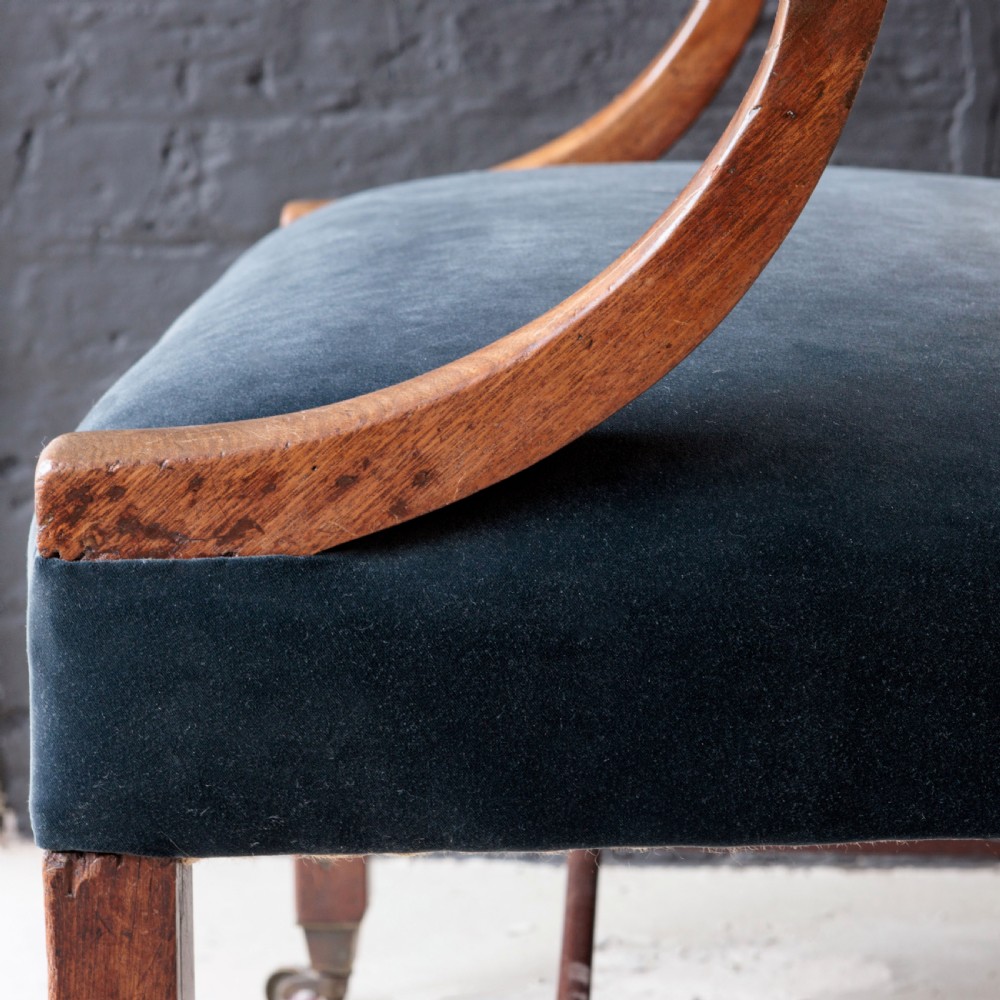 A George II Gainsborough armchair featuring an exposed timber frame, a high beige linen backrest, padded striped armrests, and a deep blue velvet upholstered seat on square legs with brass castors. View X