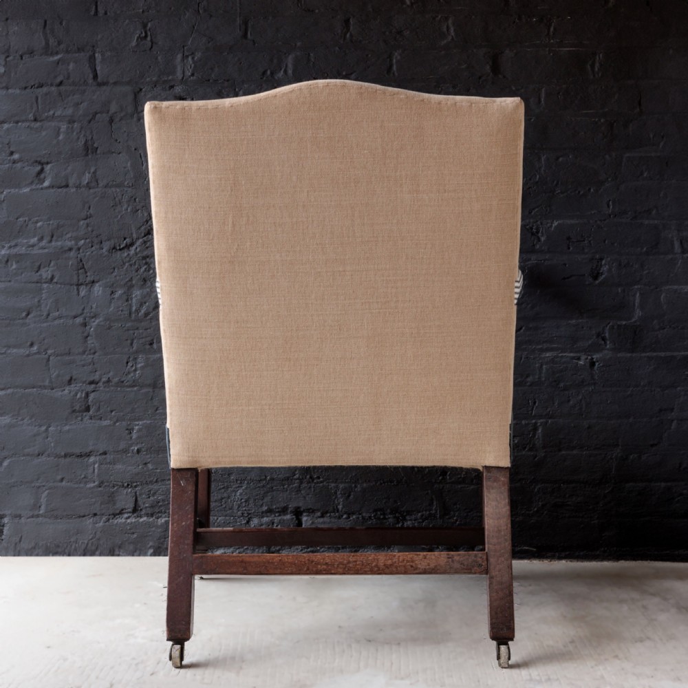 A George II Gainsborough armchair featuring an exposed timber frame, a high beige linen backrest, padded striped armrests, and a deep blue velvet upholstered seat on square legs with brass castors. View XI