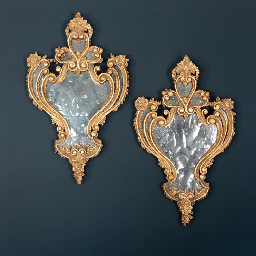 A pair of 18th-century Italian giltwood wall mirrors featuring carved scrollwork and foxed mirror plates.
