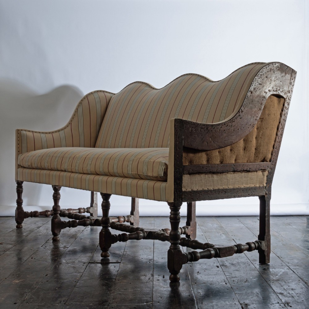 A 17th century wingback sofa featuring striped upholstery, exposed jute side panels, turned wooden legs, and stretchers.