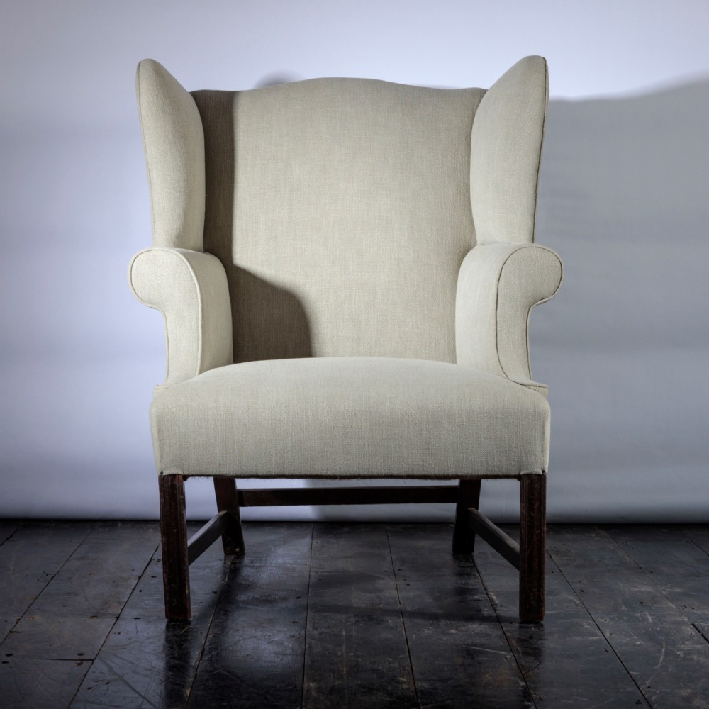 An 18th-century Georgian style wingback armchair upholstered in neutral cream linen, supported by dark square timber legs with H-stretchers.