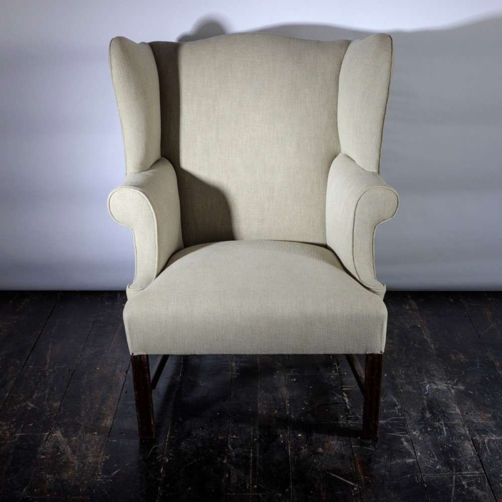 An 18th-century Georgian style wingback armchair upholstered in neutral cream linen, supported by dark square timber legs with H-stretchers. View IV