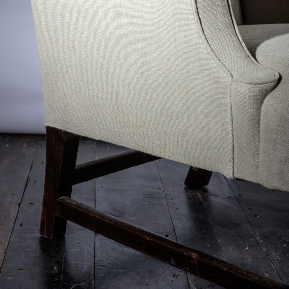 An 18th-century Georgian style wingback armchair upholstered in neutral cream linen, supported by dark square timber legs with H-stretchers. View V