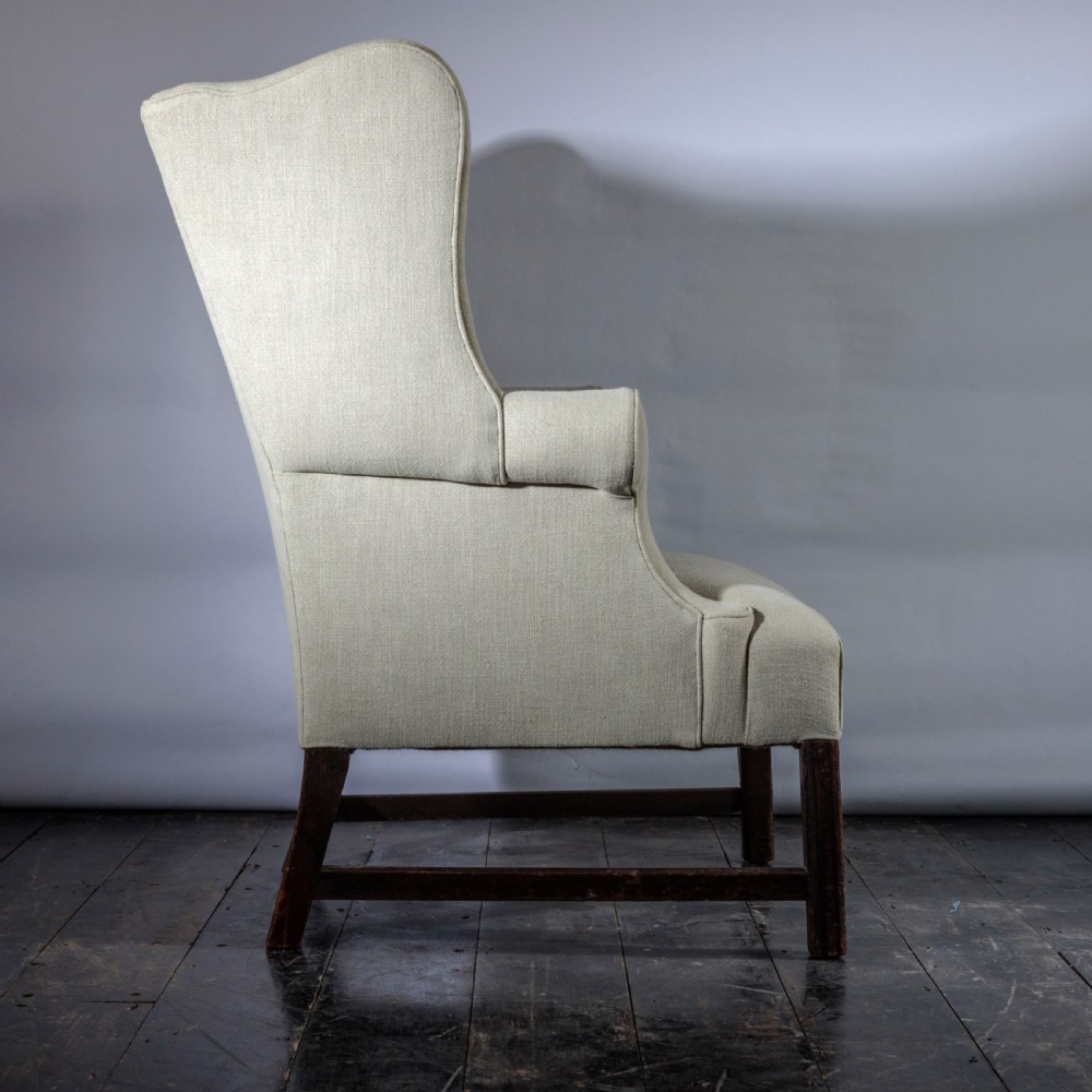 An 18th-century Georgian style wingback armchair upholstered in neutral cream linen, supported by dark square timber legs with H-stretchers. View VI
