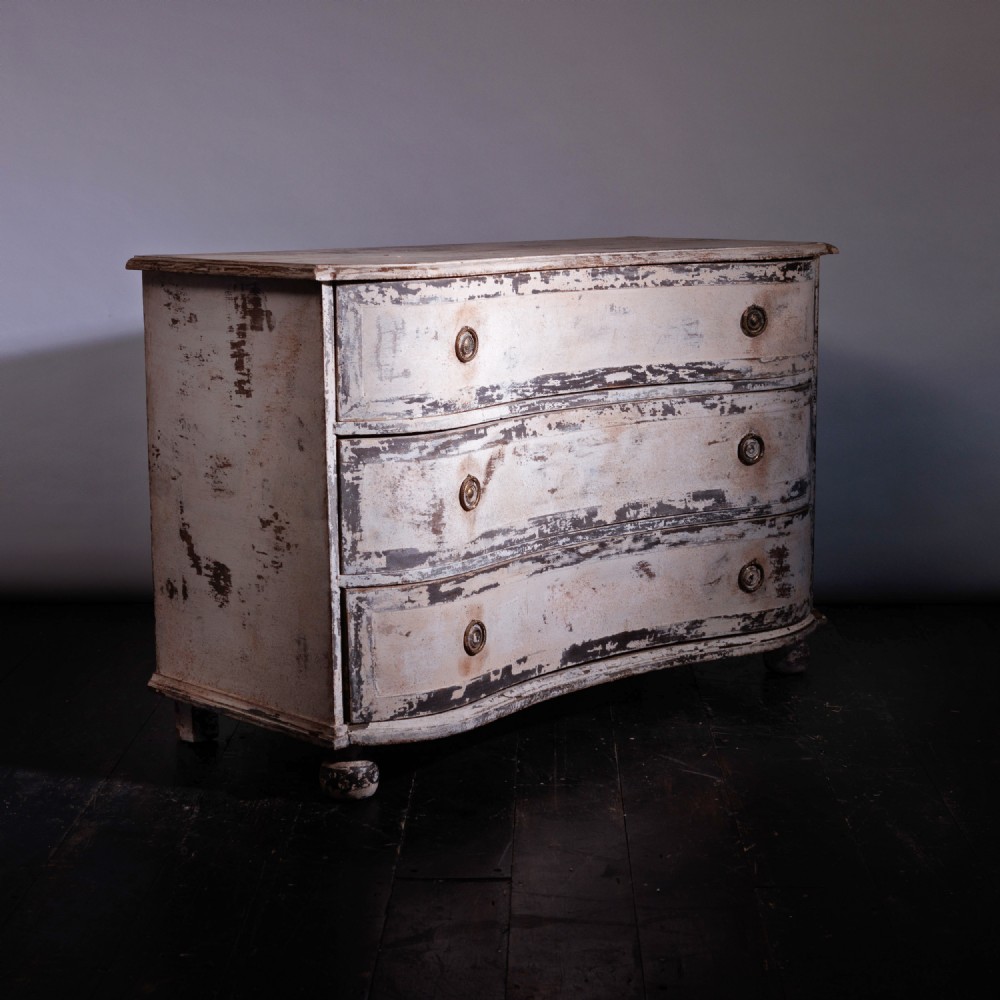An 18th-century French oak chest of drawers featuring three serpentine-fronted drawers with heavily distressed grey and white paint and circular brass ring handles.
