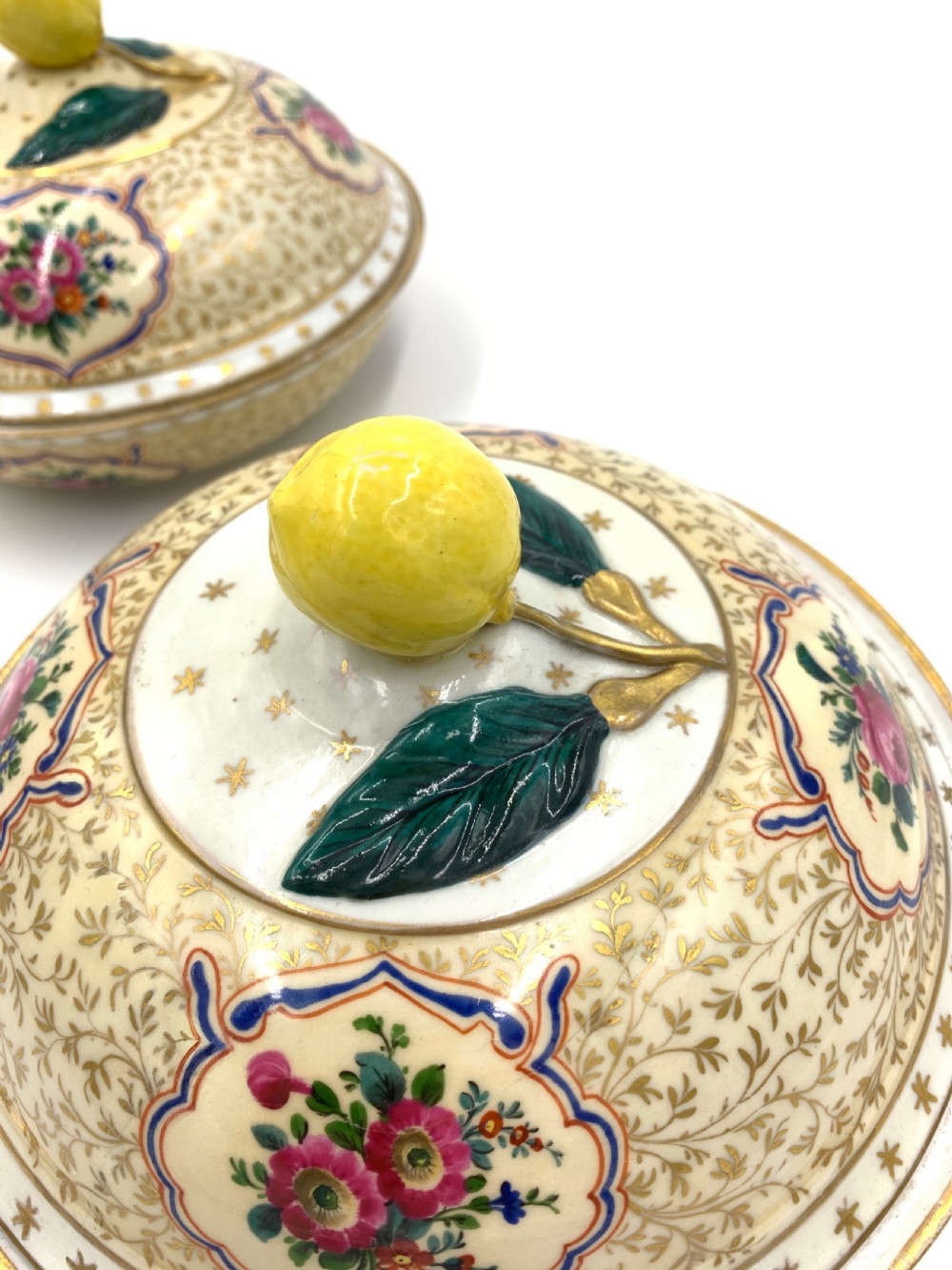 A pair of circular porcelain bonbonnieres featuring cream ground with gold leaf scrollwork, hand-painted floral cartouches, and sculpted lemon finials on the lids. View VI