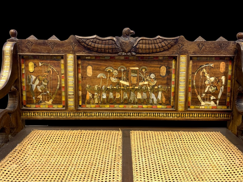 A three-piece nineteenth-century Egyptian Revival furniture suite consisting of a settee and two armchairs with carved winged crestings, figural marquetry backrests, and cane seats. View XIX