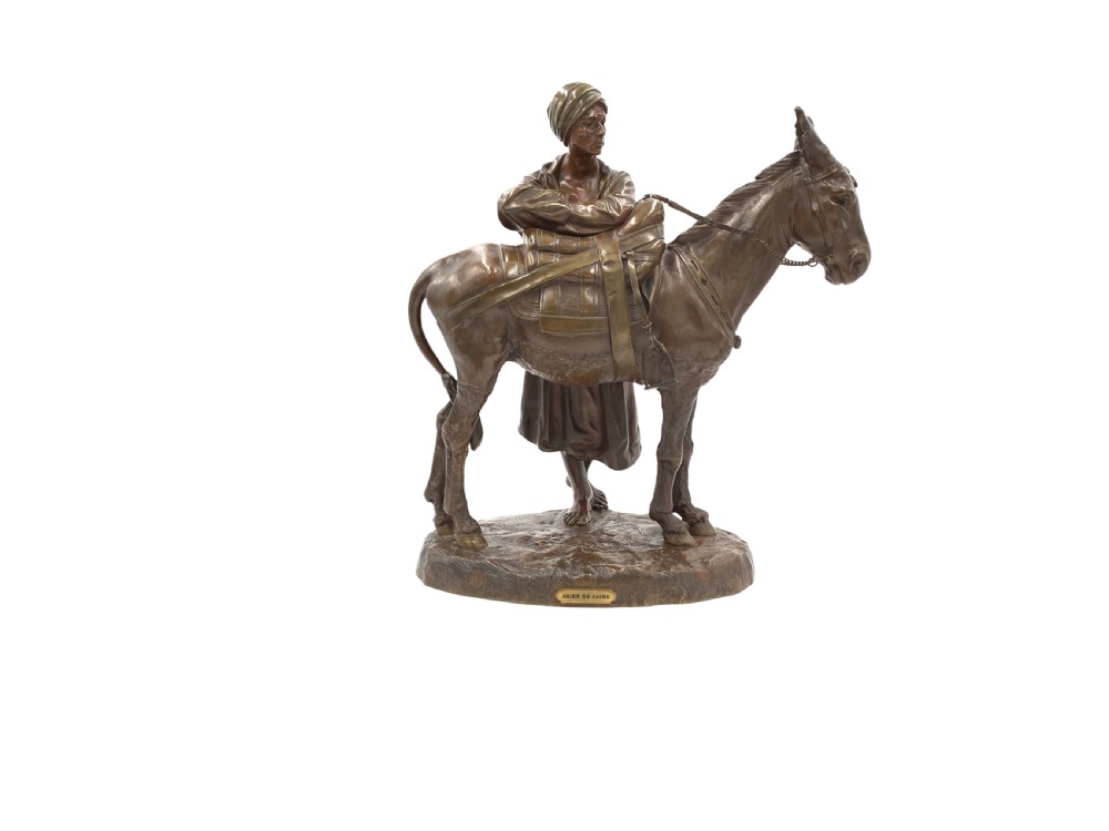A dark patinated bronze sculpture featuring a man in traditional North African attire leaning against a saddled donkey on a naturalistic base with a title plaque.