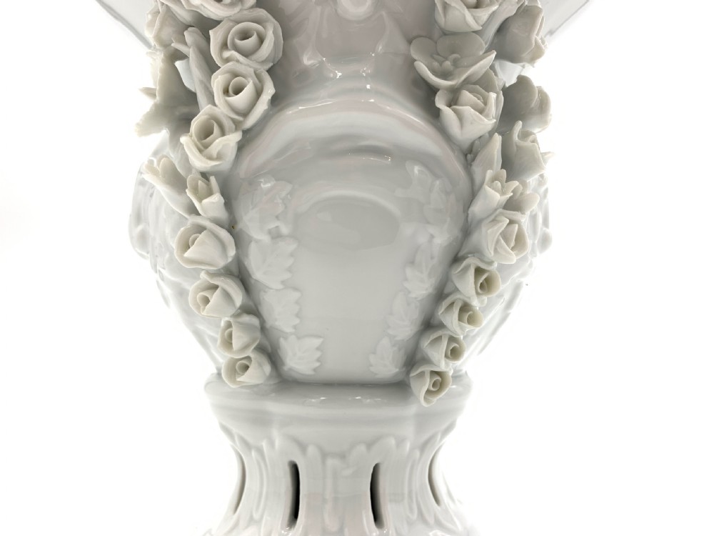 A white glazed porcelain urn featuring applied floral garlands, a pedestal base, and a pierced lid topped with a rose finial. View V